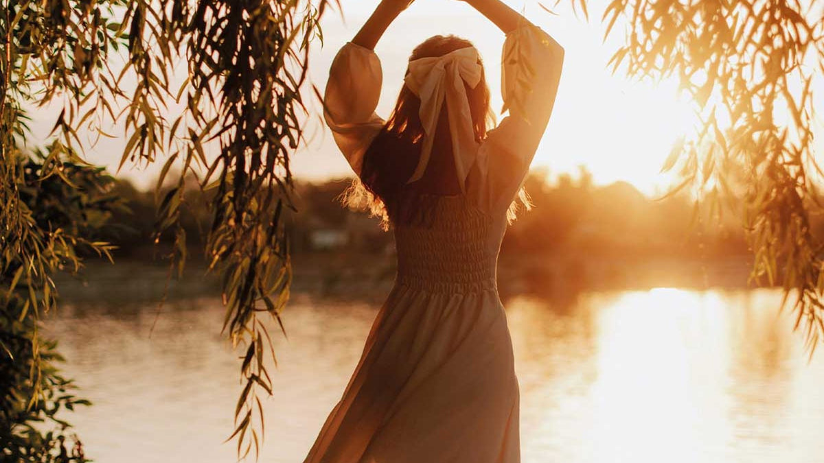 Woman in a summer dress stretching her arms up beside a lake at sunset, framed by willow branches