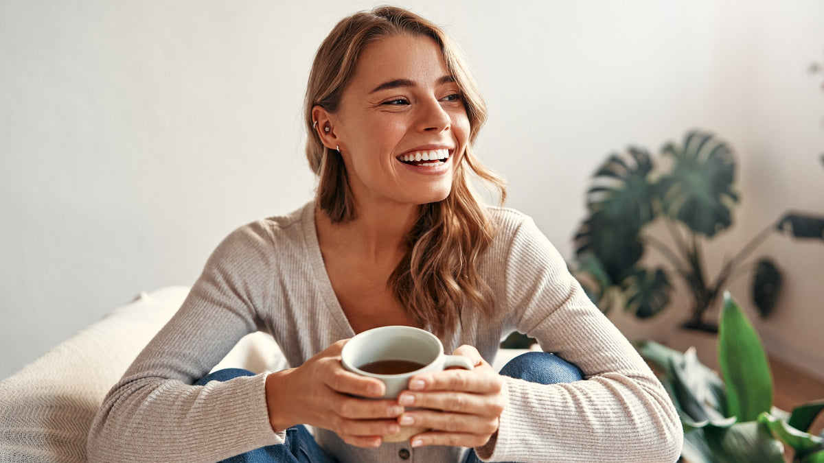 A person laughing while holding a warm mug in a bright room with plants