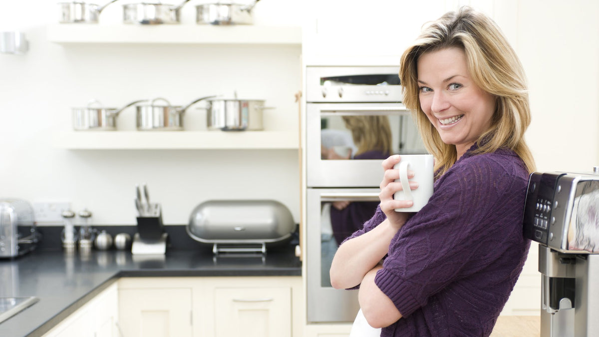 Smiling woman holding a white mug in a bright kitchen beside an espresso machine
