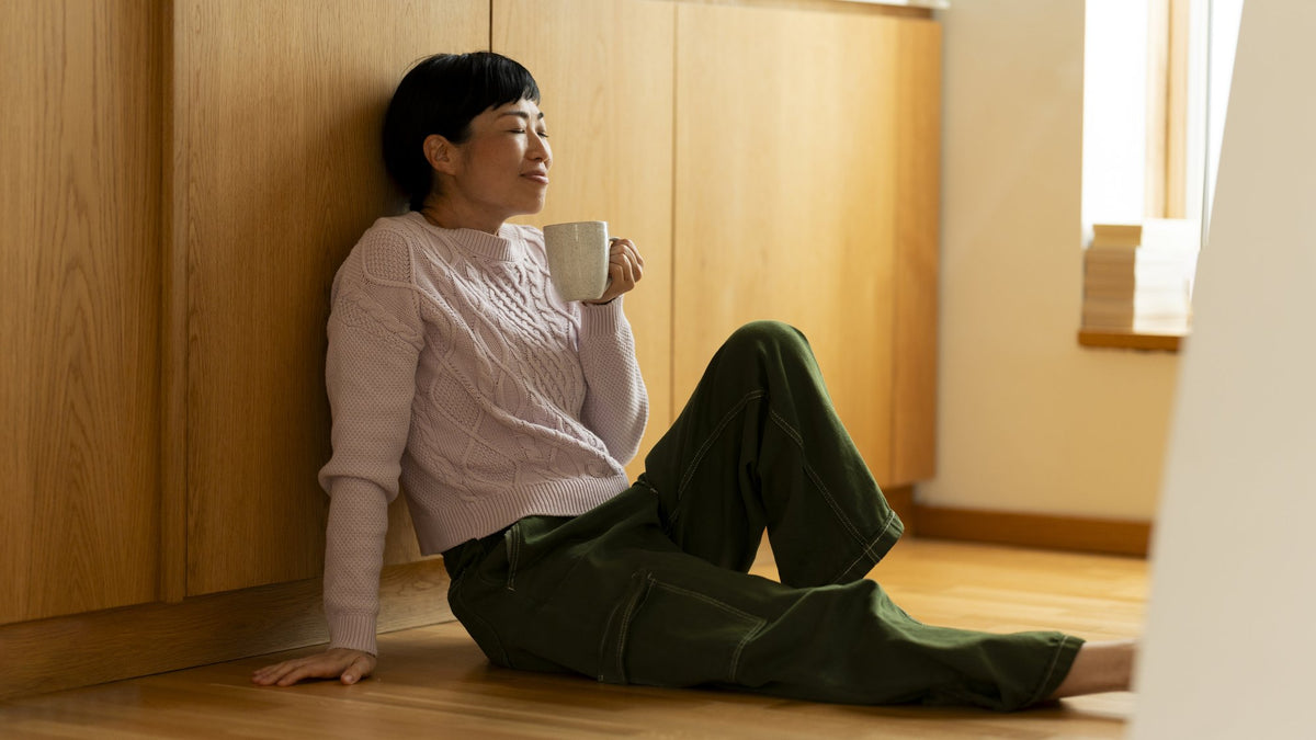 Person sitting on a wooden kitchen floor sipping from a mug in warm afternoon light