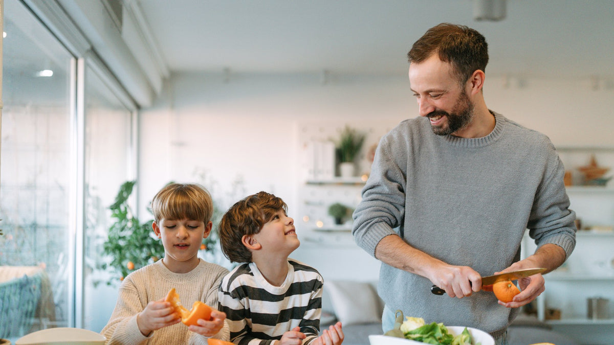 Father slicing an orange beside a salad bowl while two boys help prepare a healthy meal in a bright kitchen
