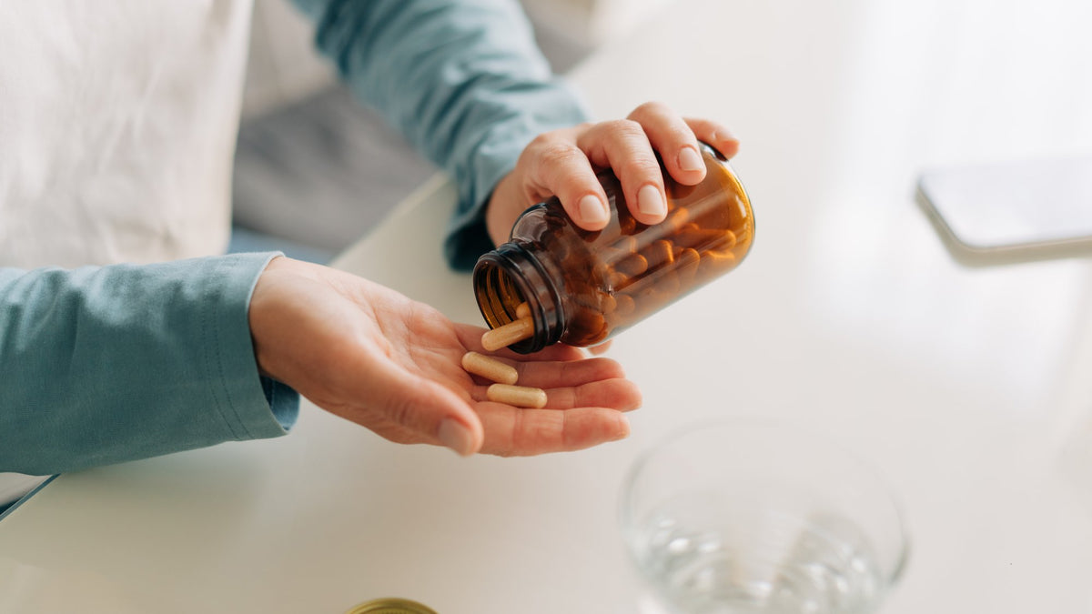 Hands pouring capsules from an amber supplement bottle into a palm beside a glass of water
