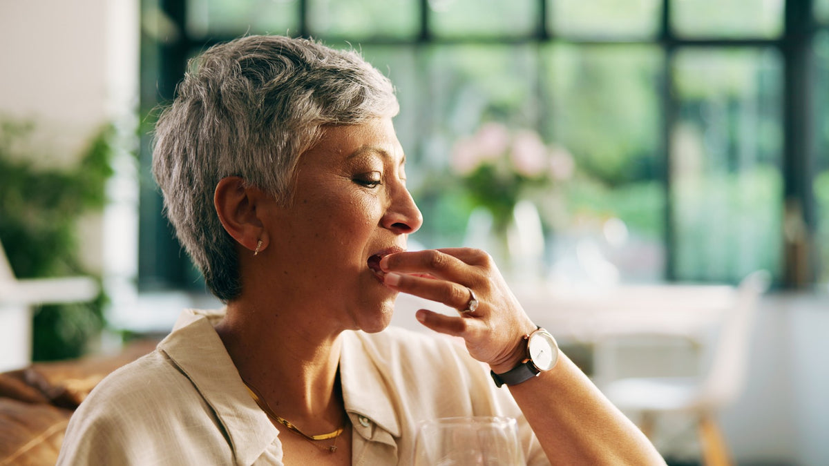 Woman with short grey hair taking a capsule with a glass of water in a bright living room