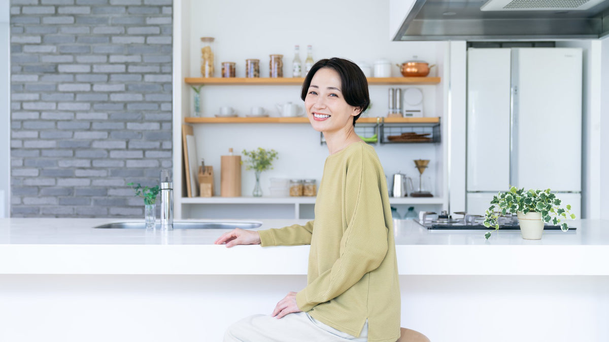 Smiling woman sitting at a bright white kitchen counter with open shelves and plants behind her