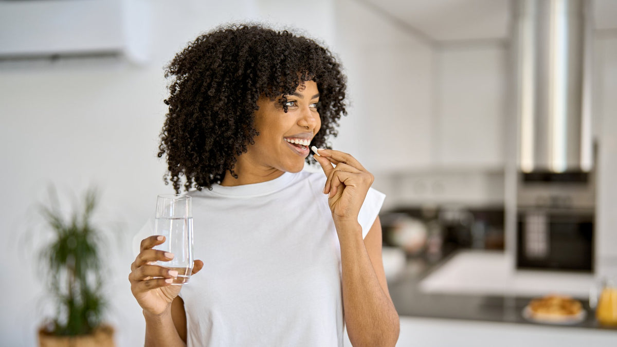 A smiling person in a bright kitchen taking a capsule with a glass of water