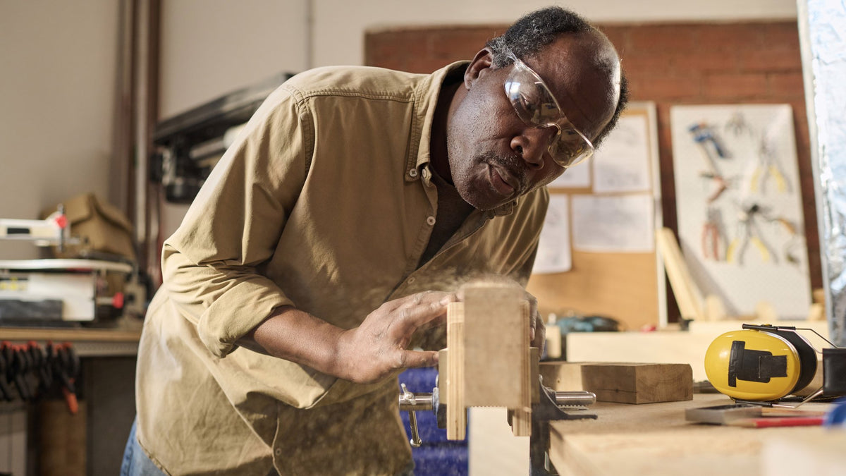 Older man in safety glasses blowing sawdust off a wooden board in his workshop