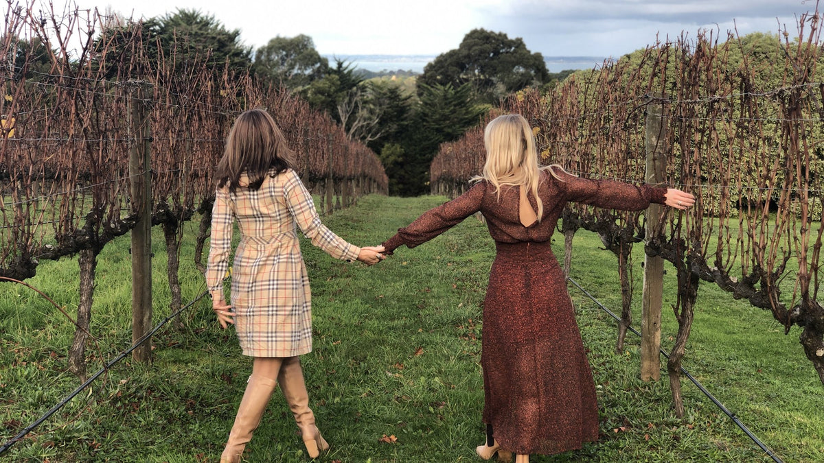 Two friends walking hand in hand between rows of vines on a grassy vineyard path