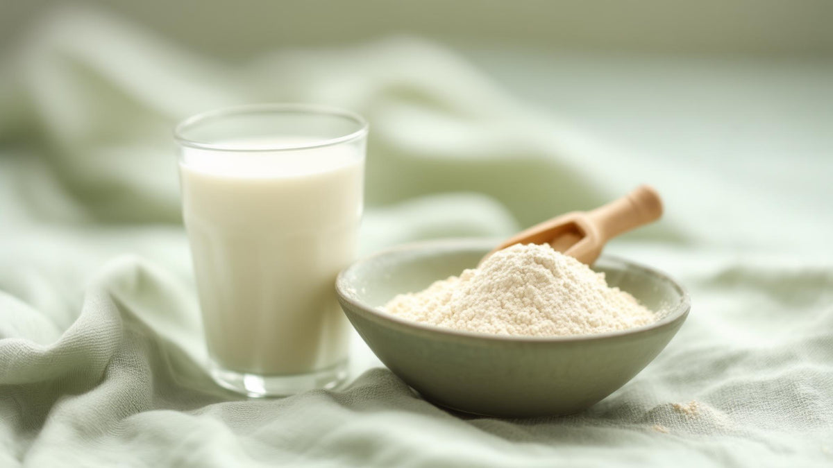 A glass of milk beside a bowl of colostrum powder with a wooden scoop on linen