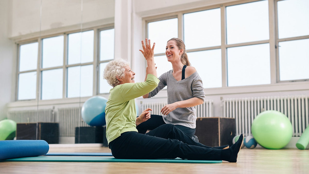 Older woman on an exercise mat giving a high five to her instructor in a bright studio
