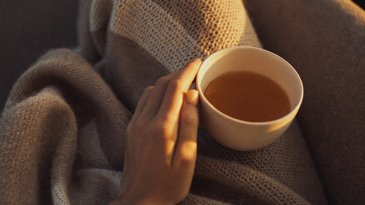 Hand holding a cup of herbal tea against a soft knit blanket in warm evening light