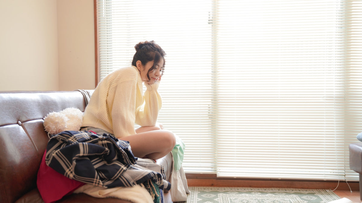 A person on a sofa with one cheek resting on a hand, eyes lowered, beside a pile of laundry by a bright window