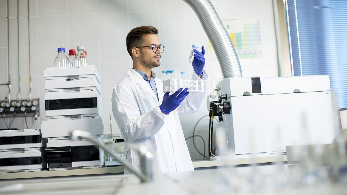 A lab researcher in a white coat and gloves holding sample vials beside testing equipment