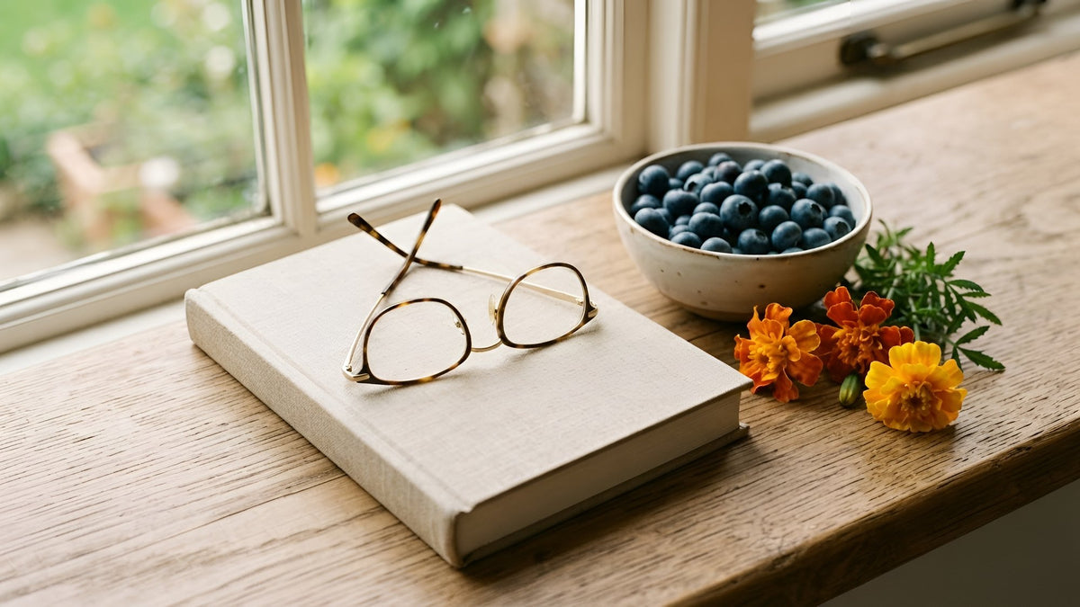 Reading glasses on a closed book beside blueberries and marigold flowers in window light