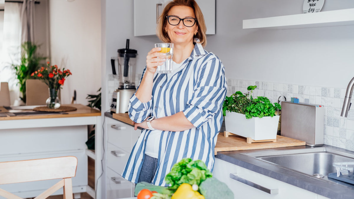 Smiling woman holding a glass of water in a bright kitchen, with leafy greens and fresh vegetables on the counter
