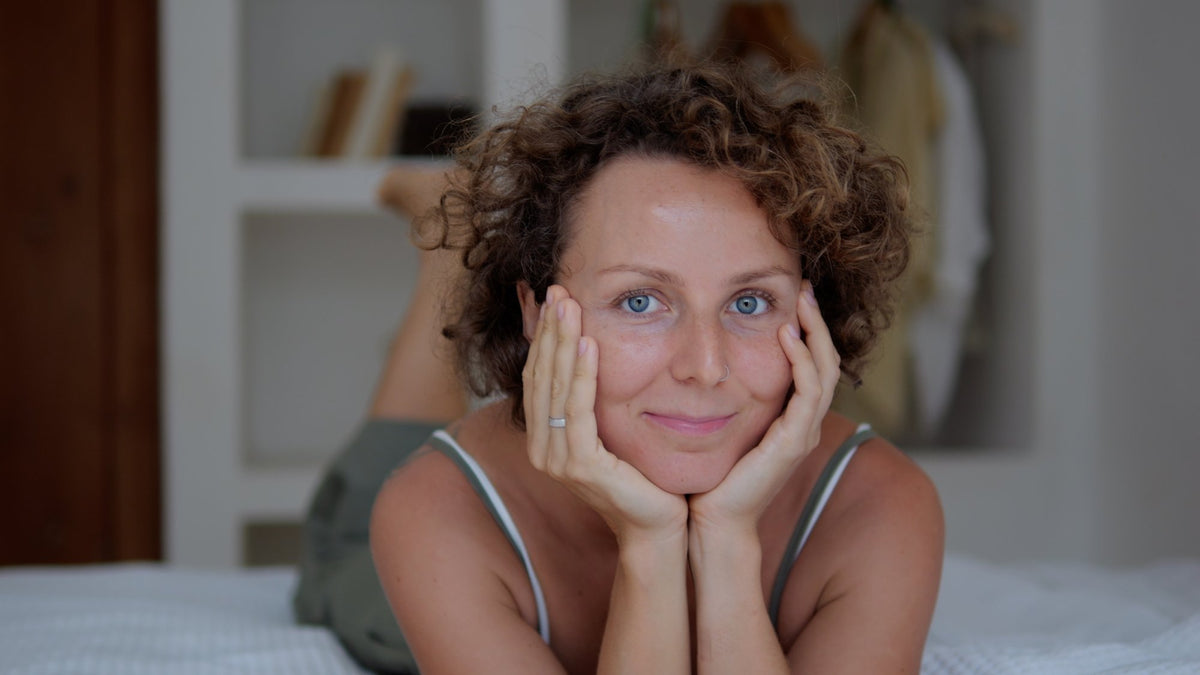 Woman in her 40s resting on a bed, chin on her hands, soft smile in natural light
