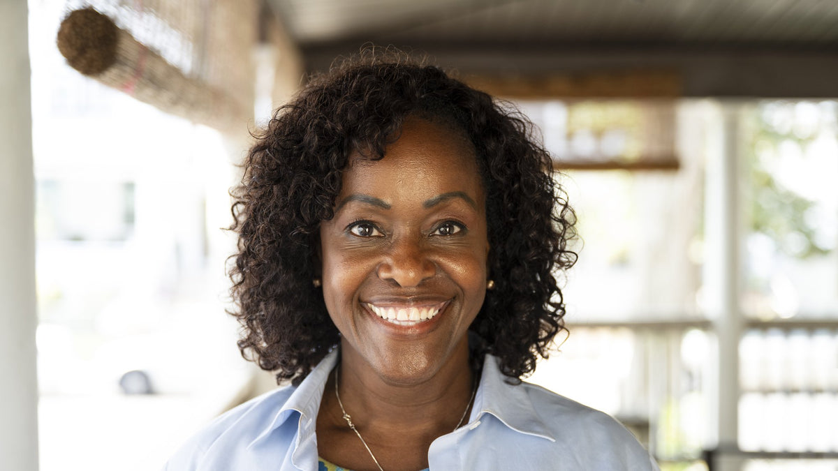 Smiling woman with curly dark hair standing on a sunlit covered porch in a light blue shirt