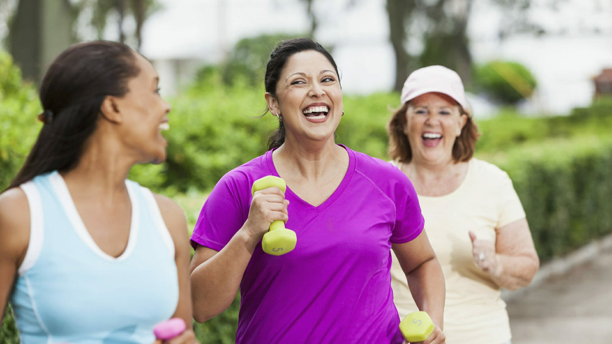 Three women laughing as they walk outdoors together carrying small hand weights