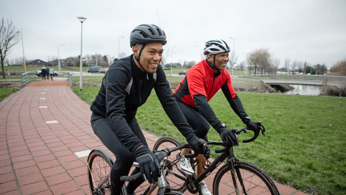 Two smiling adults in cycling jackets and helmets riding road bikes side by side on a brick path in a park