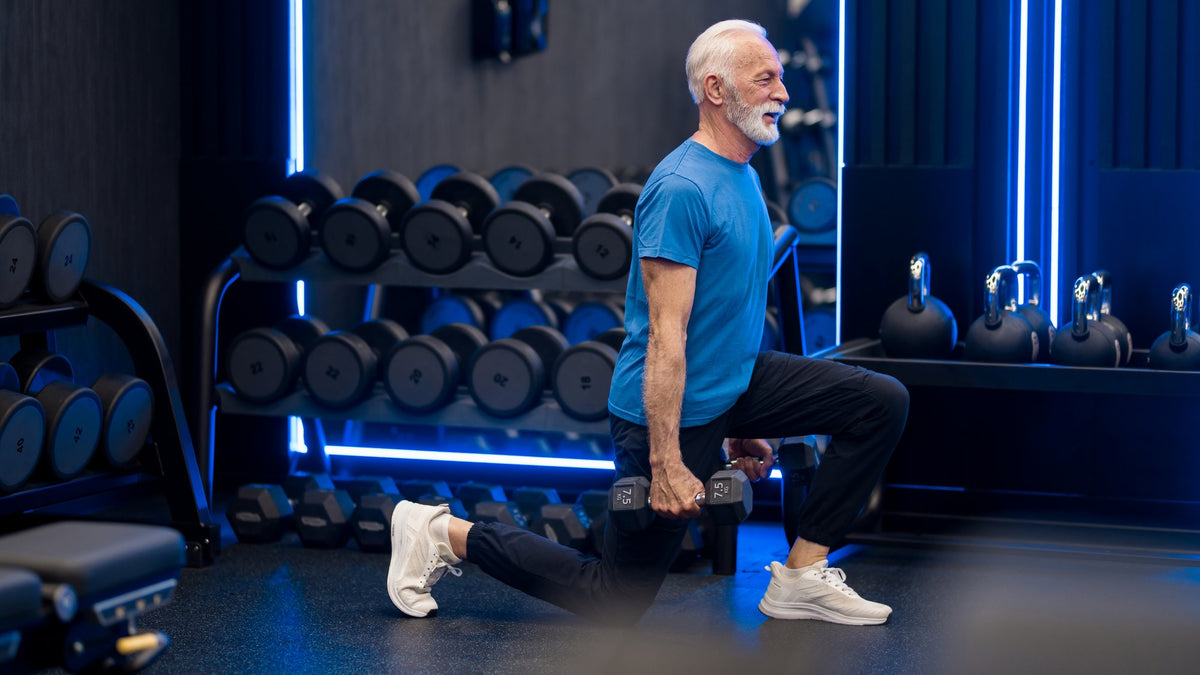 Gray-haired man in a blue T-shirt doing a dumbbell split squat in a modern gym