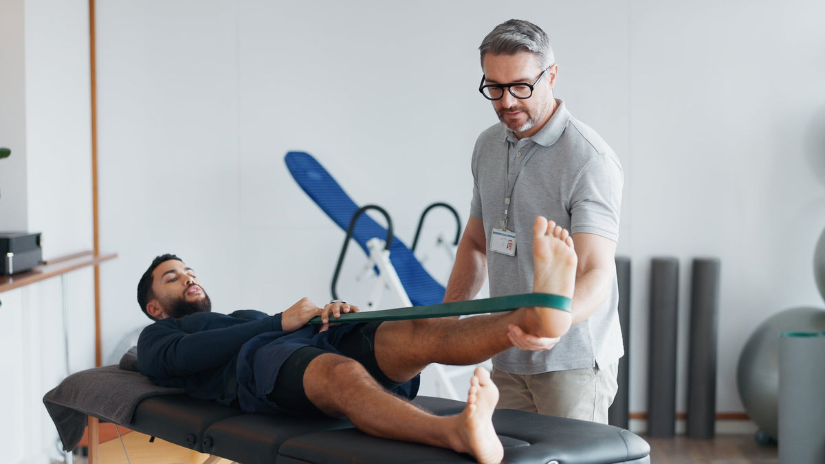 Man lying on a treatment table stretching his leg with a resistance band while a physiotherapist assists