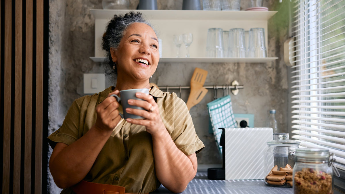 Smiling woman with grey hair holding a mug of coffee in a sunlit kitchen