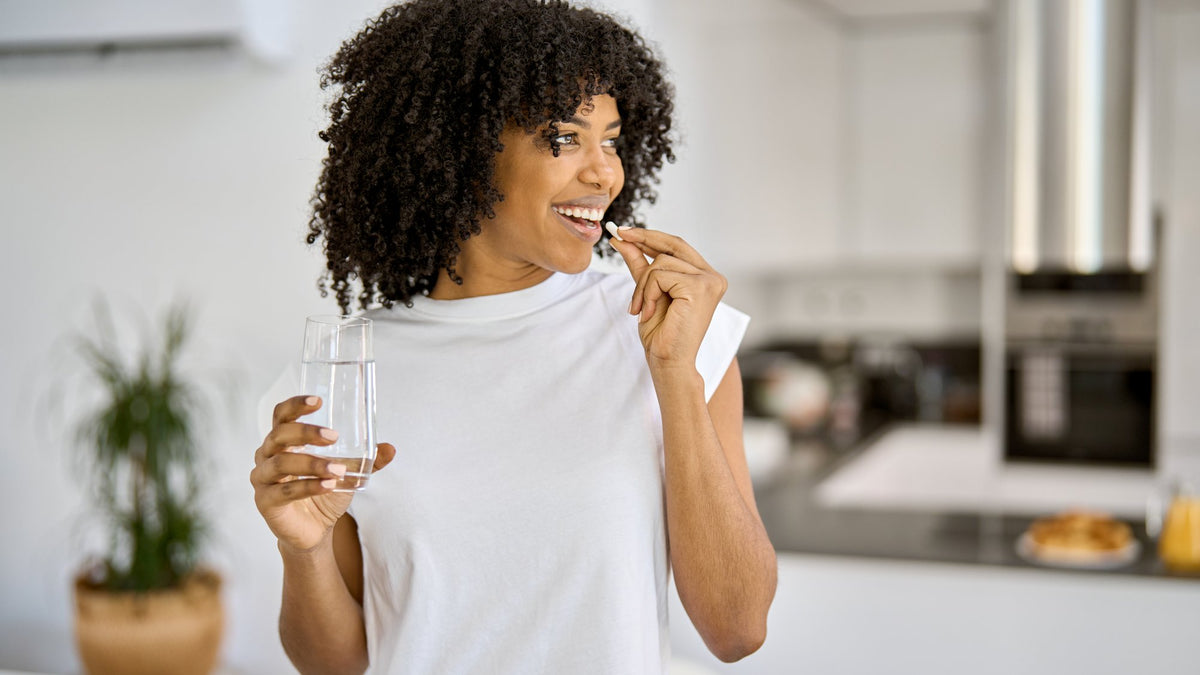 Woman in a white tee taking a capsule with a glass of water in a bright kitchen