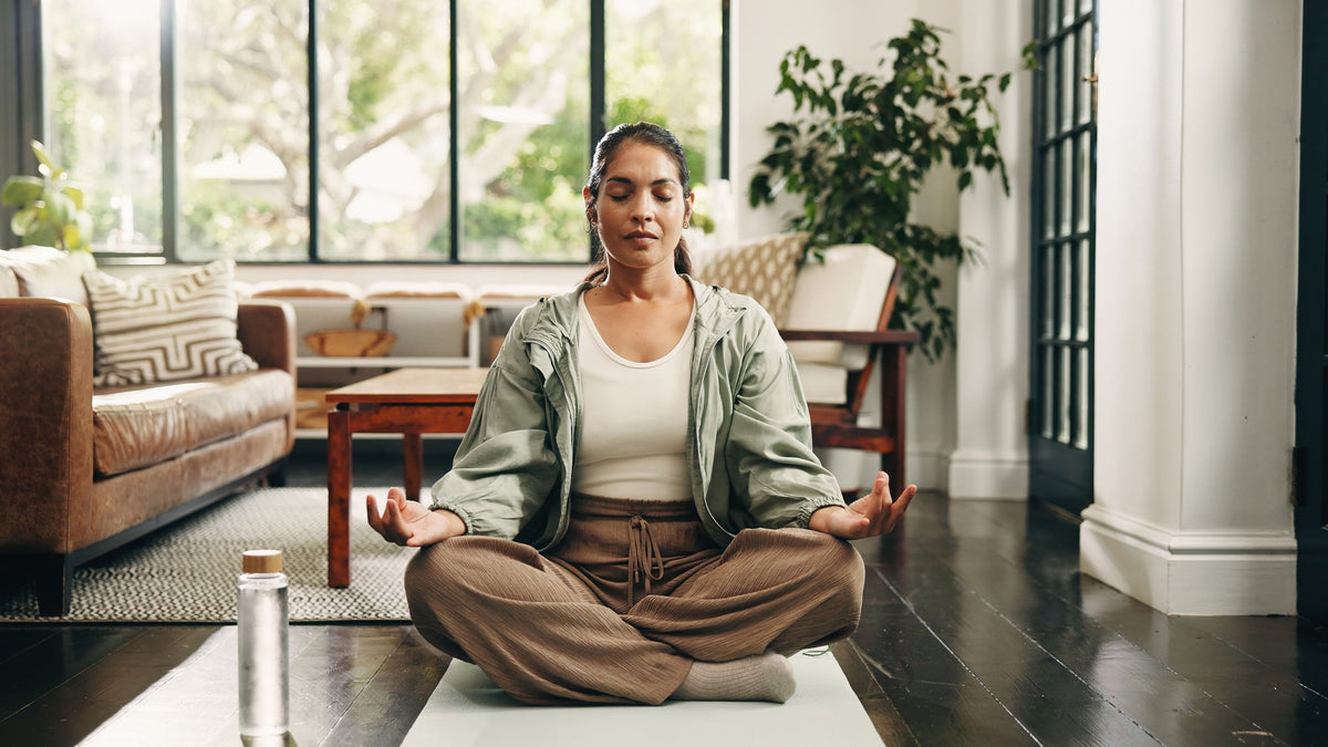 Woman sitting cross-legged on a mat with eyes closed in a bright living room