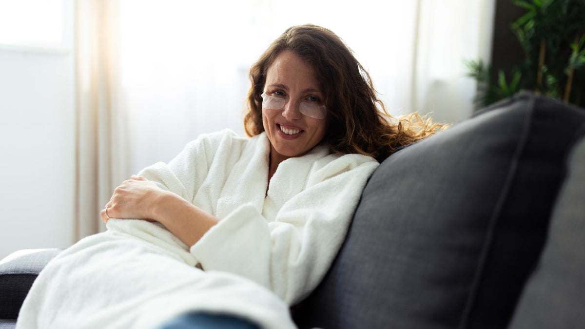 Smiling woman in a white robe and under-eye patches relaxing on a gray sofa in soft window light